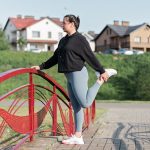 A woman in active wear performs a stretching exercise on a red bridge in a suburban park setting.