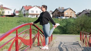 A woman in active wear performs a stretching exercise on a red bridge in a suburban park setting.